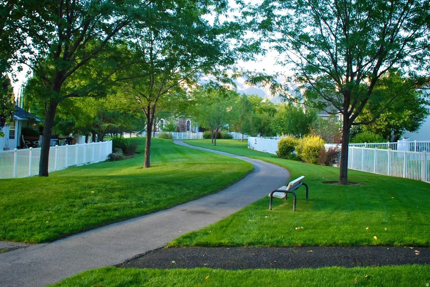 Expansive green space featuring a winding paved path, mature shade trees, and a white vinyl privacy fence