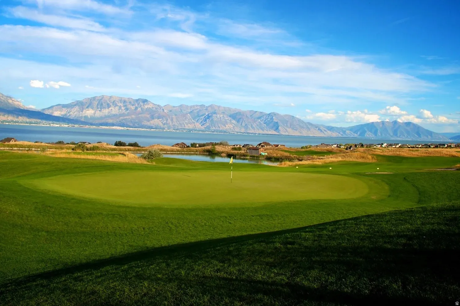 Manicured golf course green with a white flag, surrounded by a water feature and distant mountains