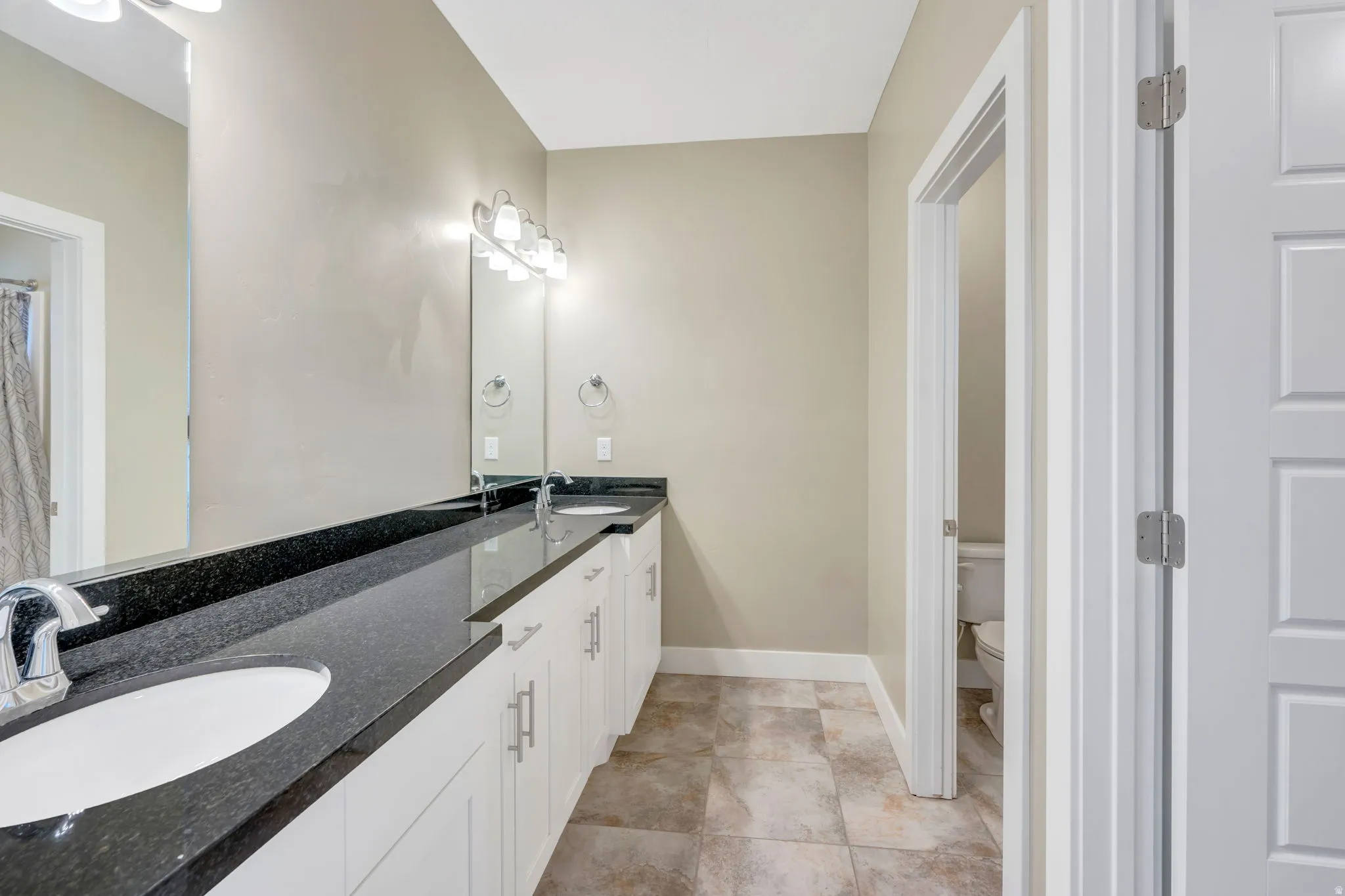 Primary bathroom featuring a long dual vanity with white cabinetry, dark stone-finish countertops, and two undermount sinks with chrome fixtures and a garden tub