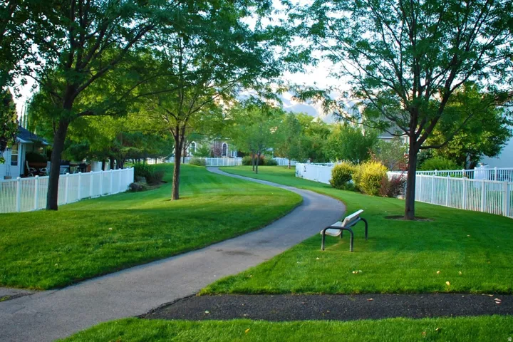 Expansive green space featuring a winding paved path, mature shade trees, and a white vinyl privacy fence