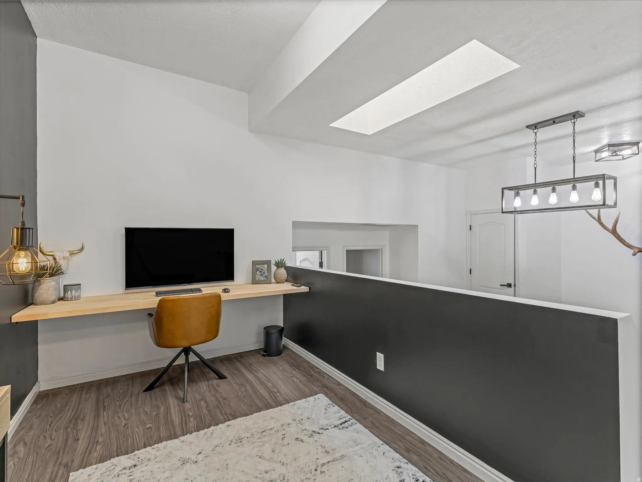 Wood-finish flooring throughout this open loft space featuring a built-in light wood desk, a skylight, and a linear pendant light fixture
