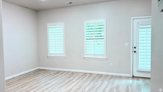 Spacious room featuring light wood-finish flooring, neutral wall paint, two windows with white plantation shutters, and a white door with an integrated shutter