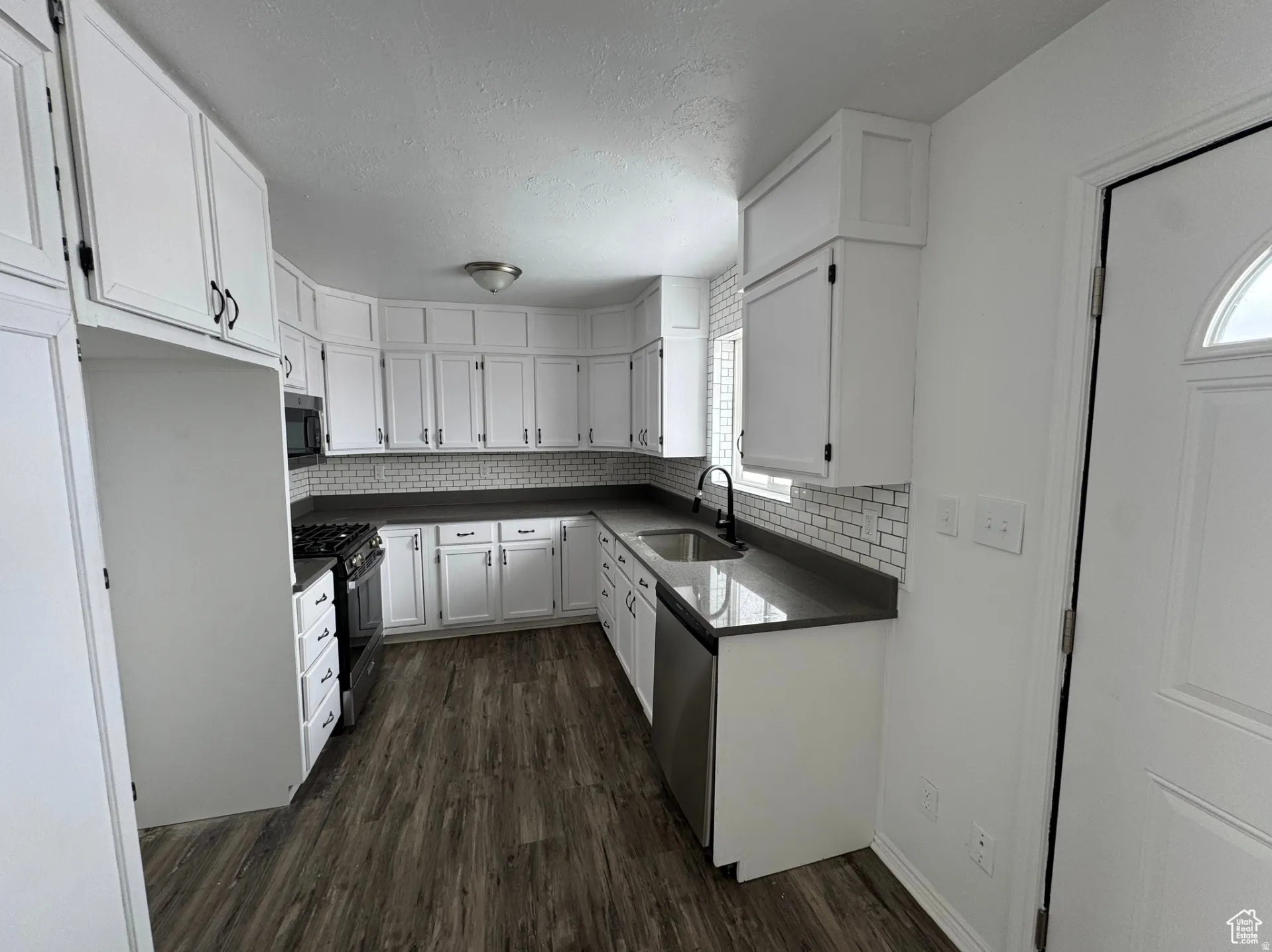 Modern kitchen featuring crisp white cabinetry with brushed nickel hardware, black countertops with subway tile backsplash, stainless steel appliances, and wood-finish flooring throughout
