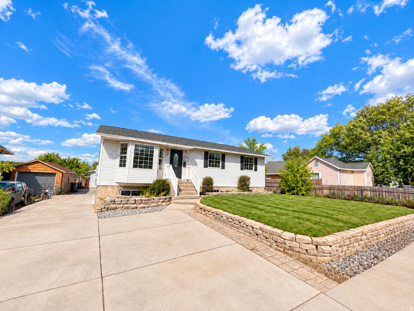 White siding exterior with black shutters, mature landscaping, stone retaining walls, and an expansive paved driveway
