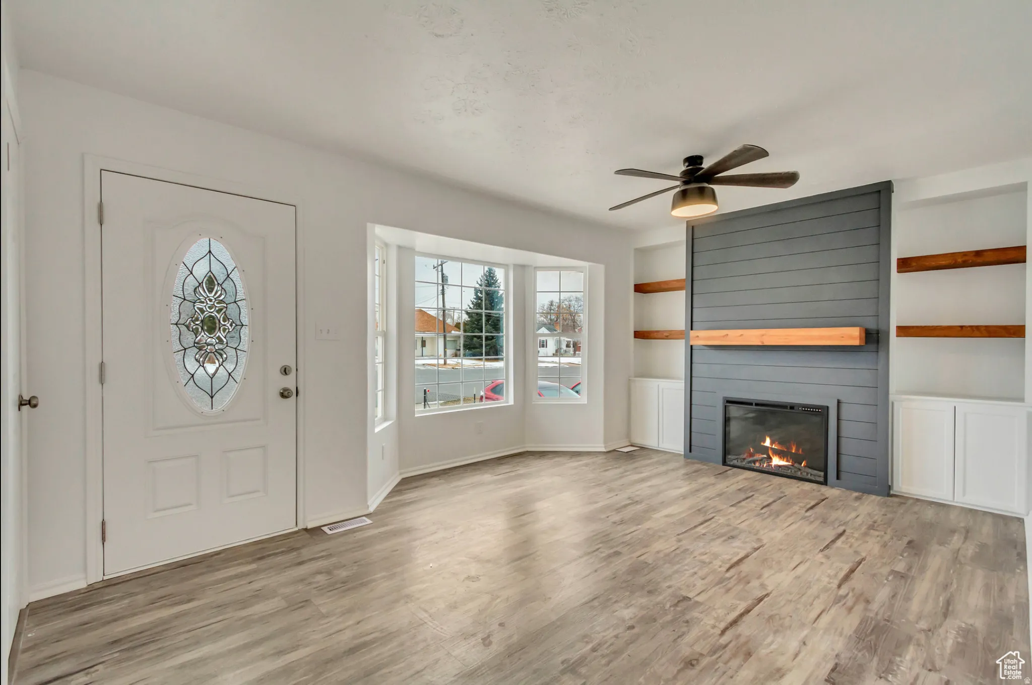 Bright and open living space featuring gleaming wood-finish flooring, a gas fireplace with gray shiplap surround and built-in shelving with wood accents, ceiling fan, and abundant natural light from French doors and windows