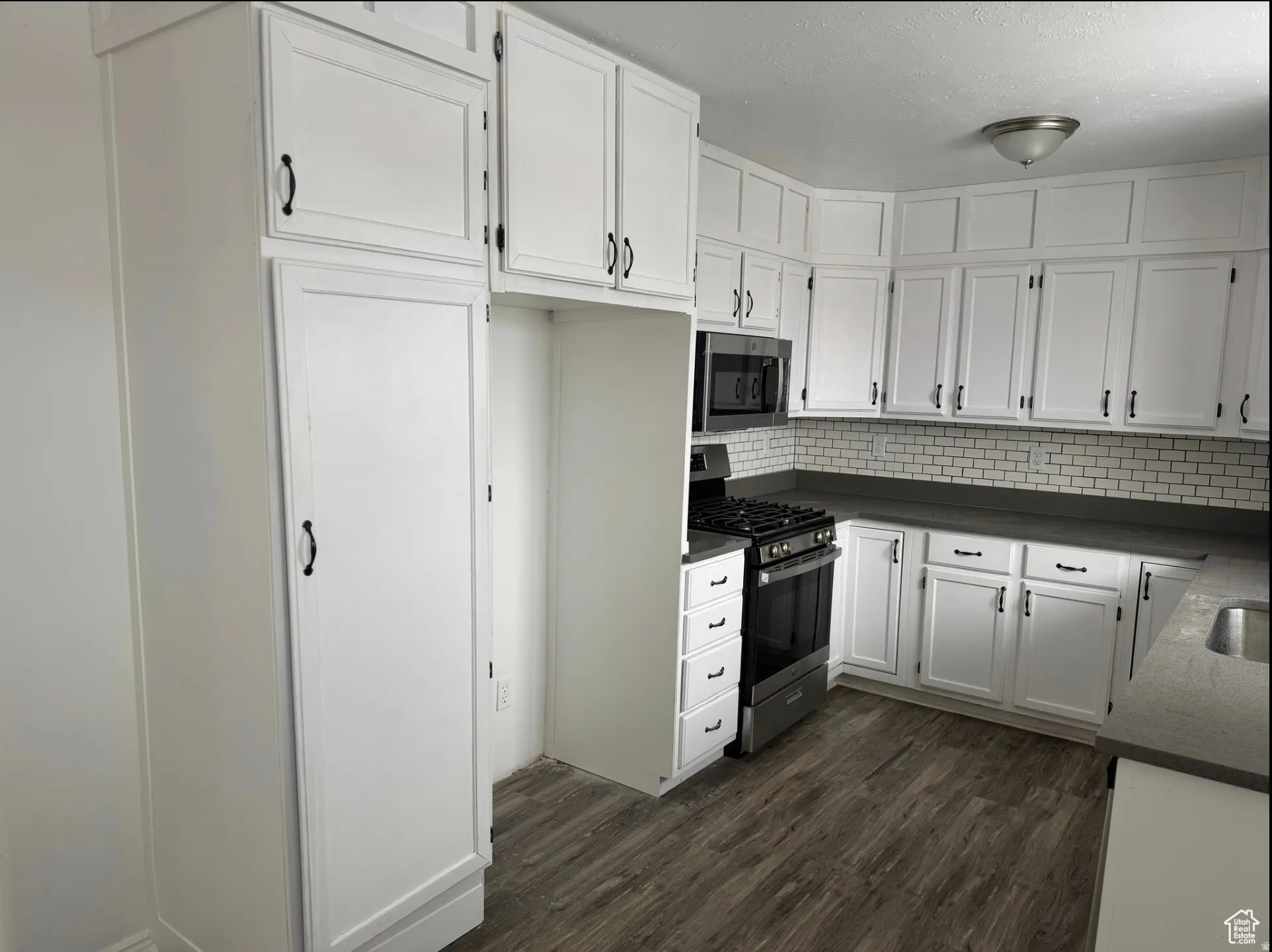 Modern kitchen featuring extensive white cabinetry with matte black hardware, dark countertops, white subway tile backsplash, and wood-finish flooring