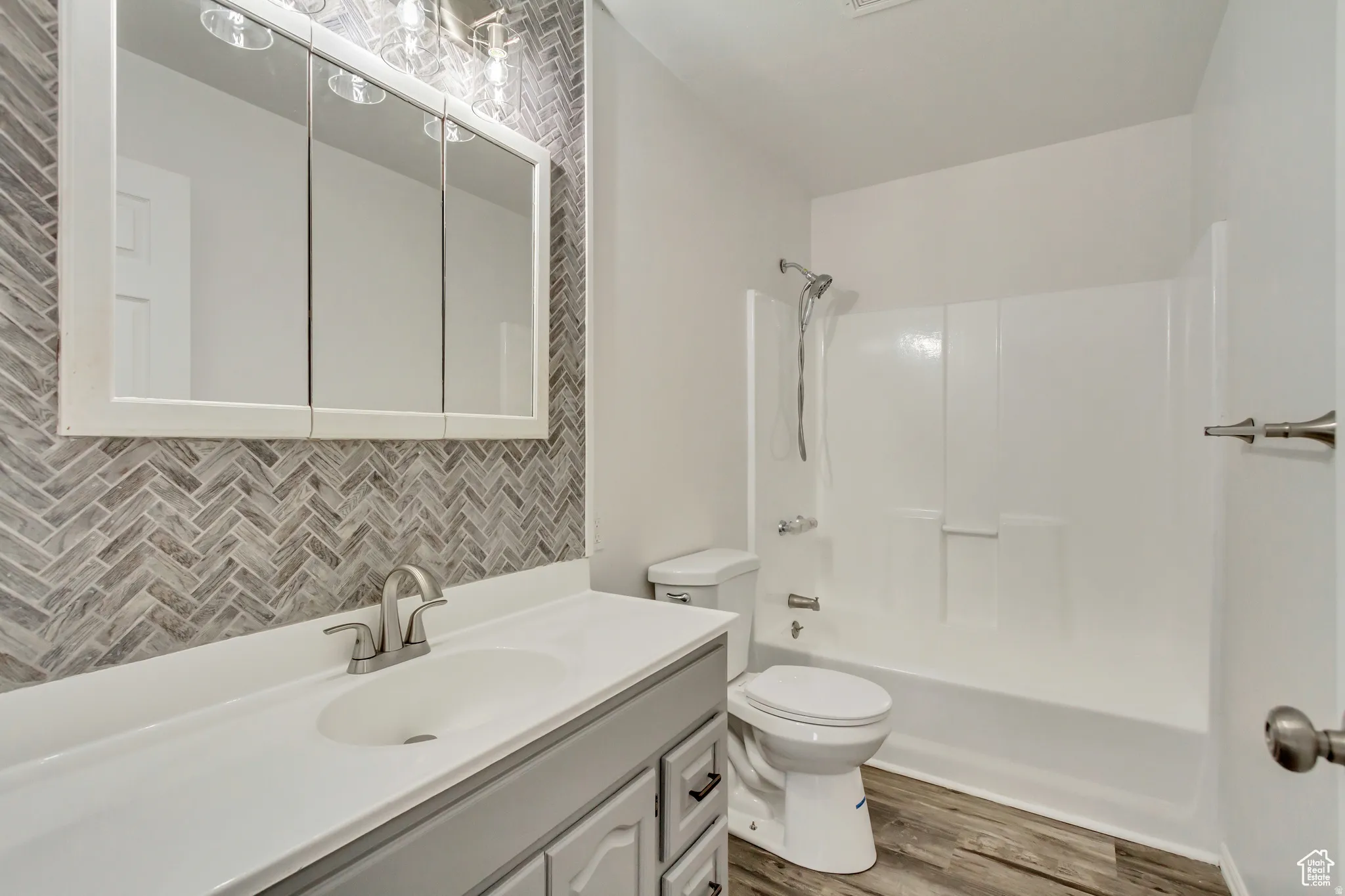 Bathroom featuring a herringbone tile accent wall, white vanity with integrated sink, brushed nickel faucet, three-door mirrored medicine cabinet, and wood-finish flooring