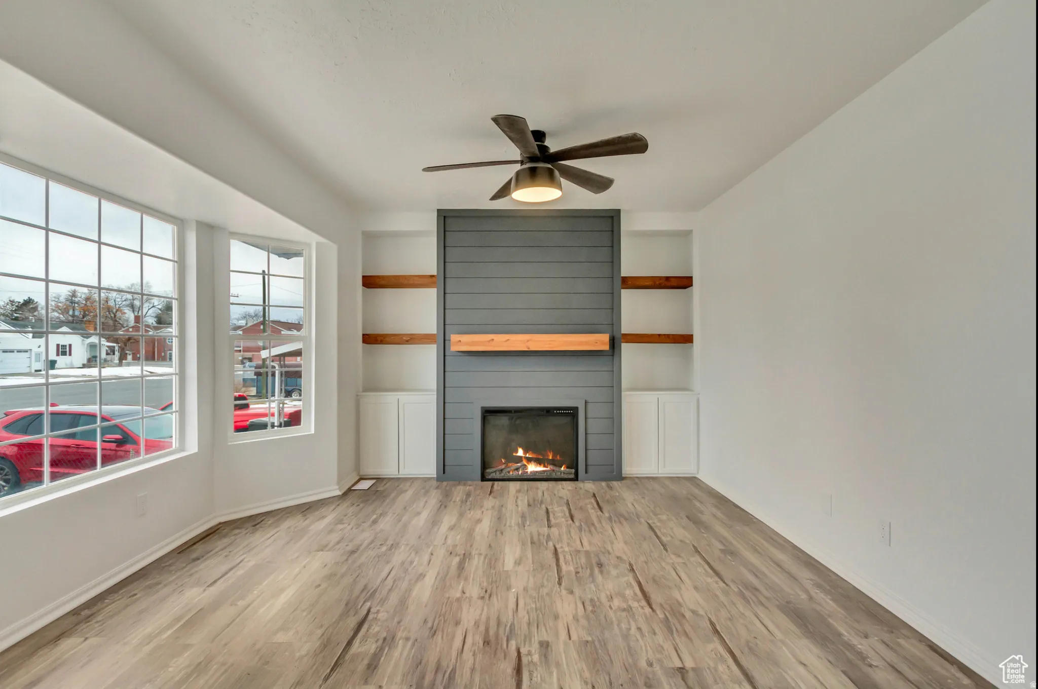 Bright, open living space featuring wood-finish flooring, a ceiling fan with integrated lighting, and a striking focal-point fireplace with horizontal shiplap detailing and warm accent lighting