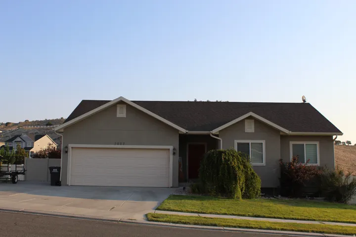 Ranch-style residence featuring a taupe stucco exterior, a dark shingle roof, and a two-car garage with a light-colored door