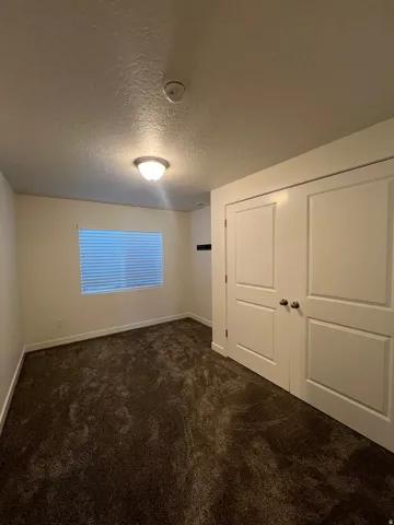 White walls, dark textured carpeting, flush-mount ceiling light, white window blinds, and white bi-fold closet doors
