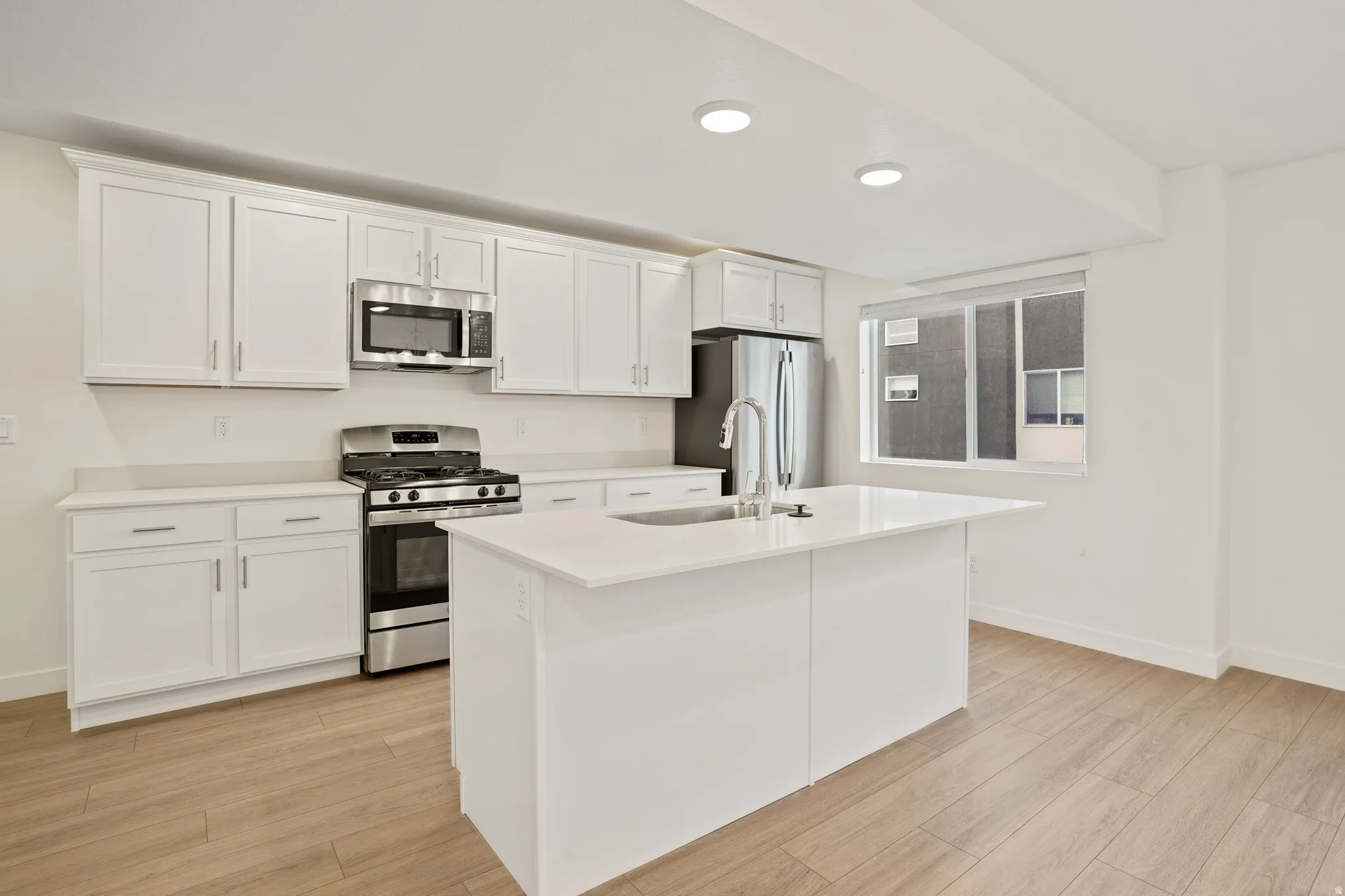 Kitchen featuring white cabinetry, stainless steel appliances, a center island with sink, light countertops, and wood-finish flooring