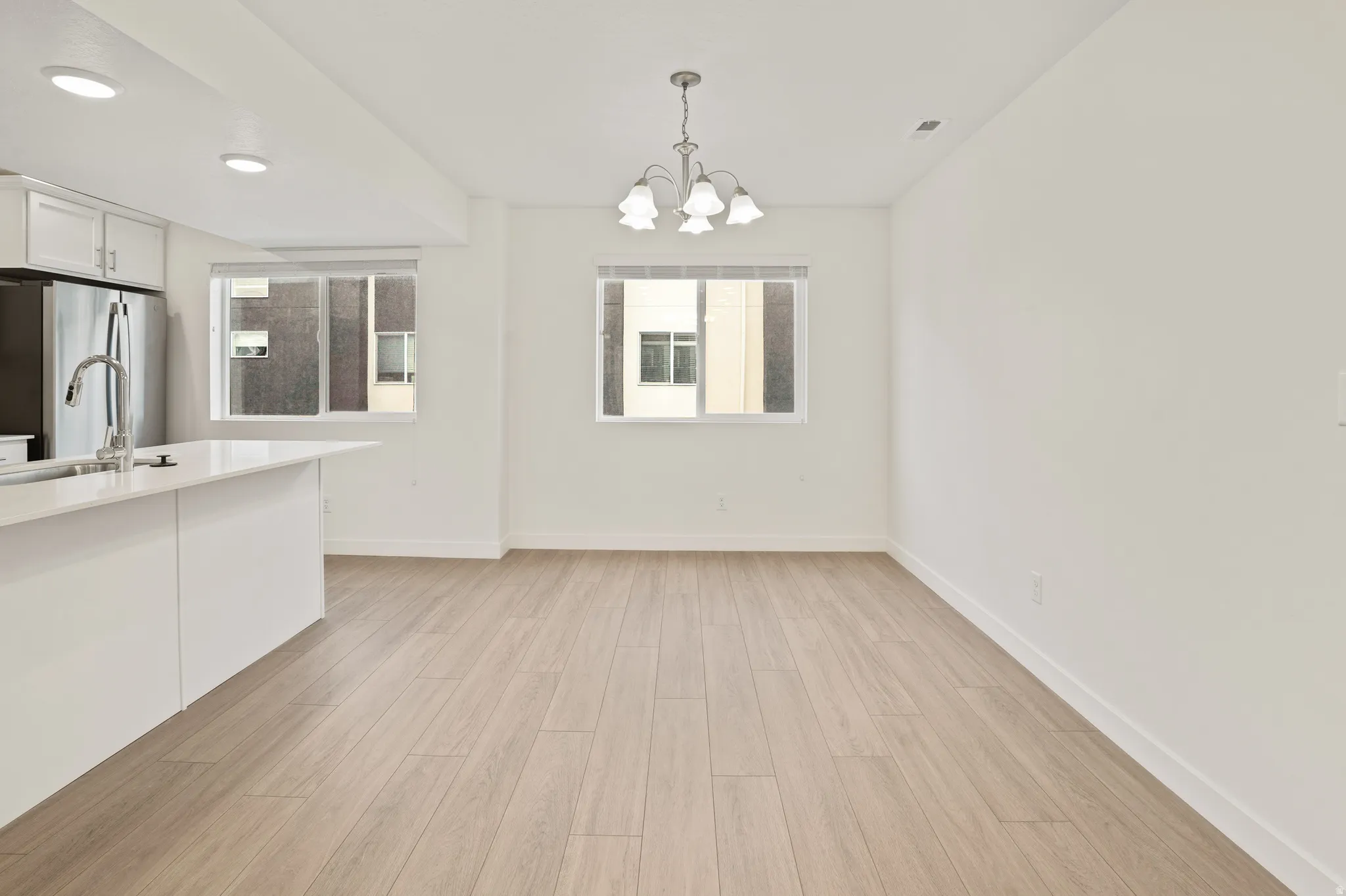 Open-concept kitchen and dining area featuring white cabinetry with integrated appliances, a sleek countertop with undermount sink, wood-finish flooring throughout, recessed ceiling lighting, and large windows providing natural light