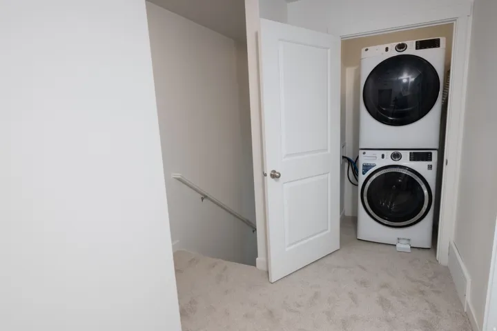 Dedicated laundry room featuring stacked washer and dryer units with modern controls, neutral carpet flooring, and ample storage cabinetry