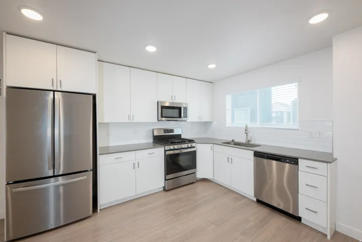 Modern kitchen featuring light wood-finish flooring, white cabinetry with silver hardware, gray countertops, a subway tile backsplash, and stainless steel appliances