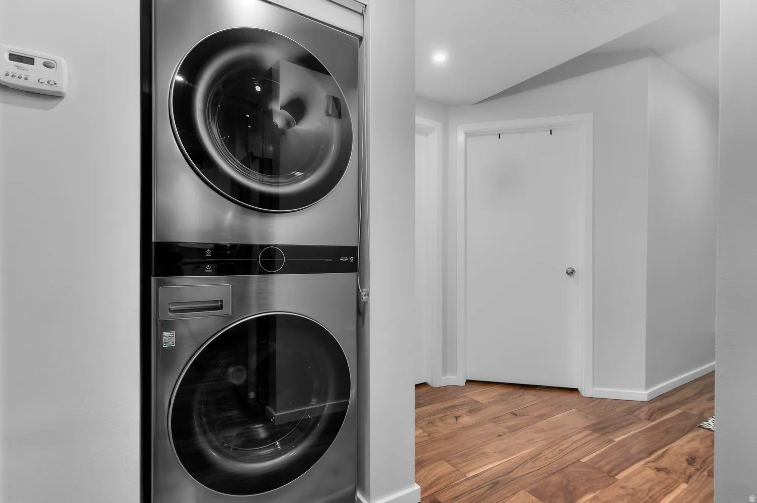 Stainless steel stackable washer and dryer unit, wood-finish flooring, white walls, white trim, and a white interior door with a circular silver handle