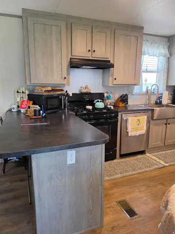 Kitchen featuring light gray wood-finish cabinetry, dark countertops, a black range with an overhead hood, a stainless steel dishwasher, and a farmhouse-style sink