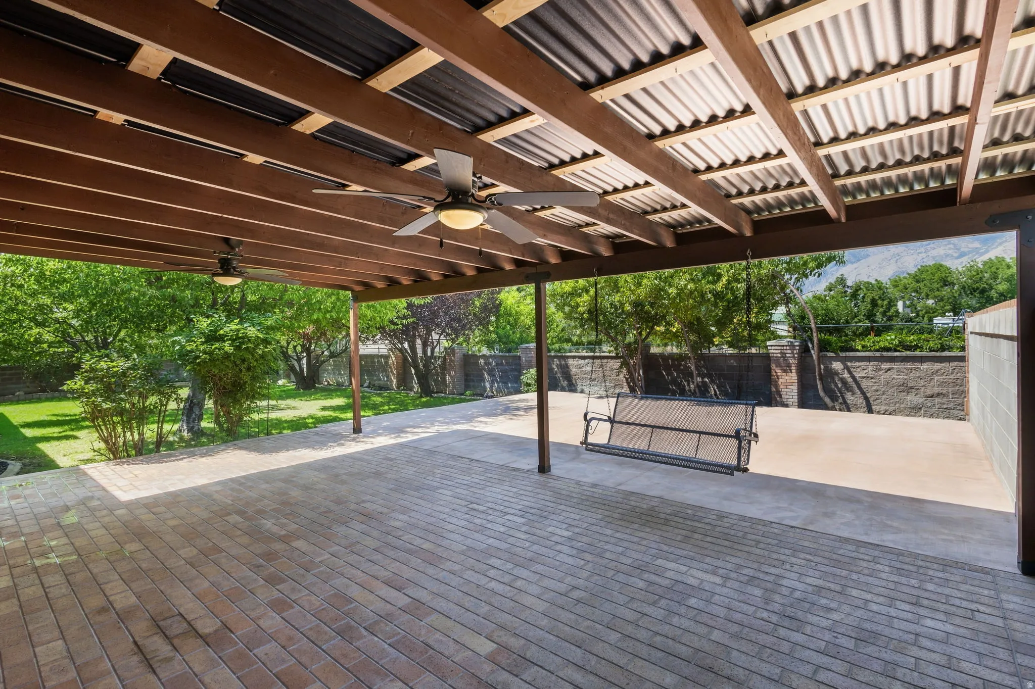 Covered patio featuring a wood beam and corrugated metal roof, two ceiling fans with integrated lighting, and brick paver flooring
