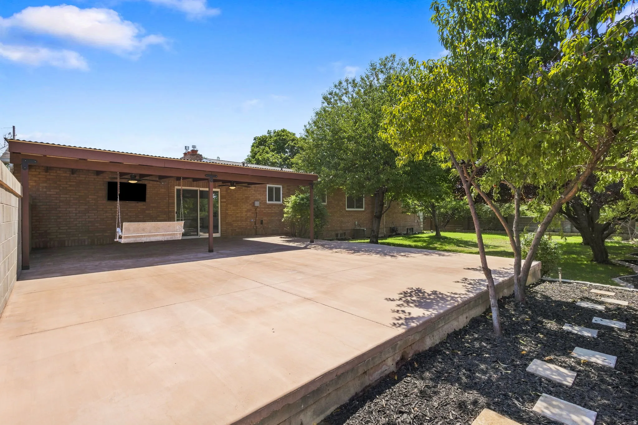 Expansive concrete patio with a covered outdoor area, brick exterior, lush green lawn, and fruit trees.