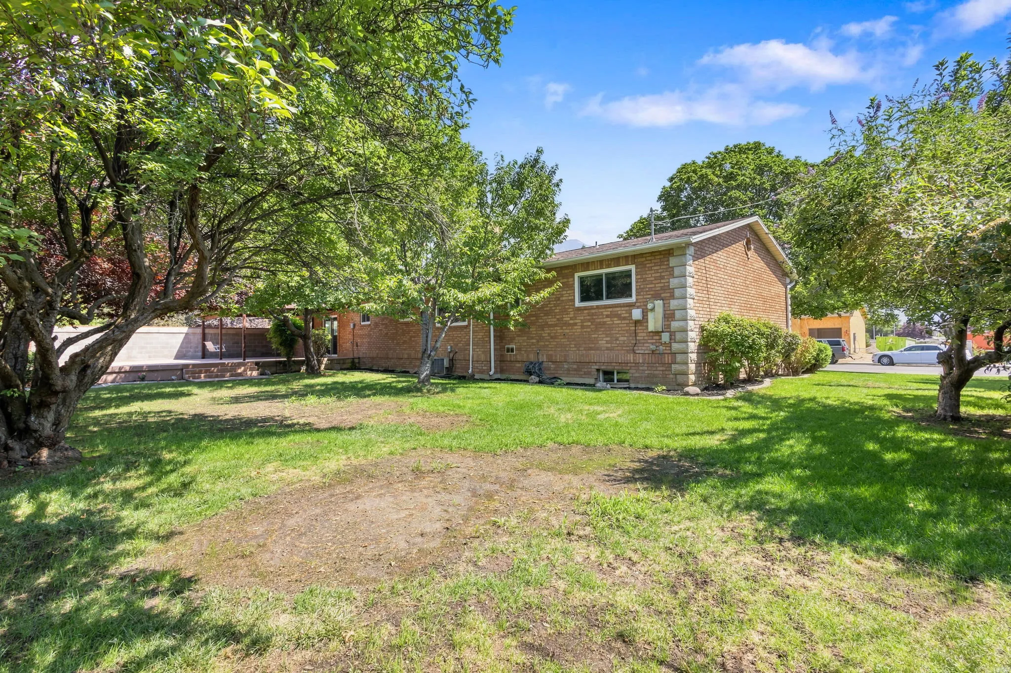 Expansive concrete patio with a covered outdoor area, brick exterior, lush green lawn, and fruit trees.