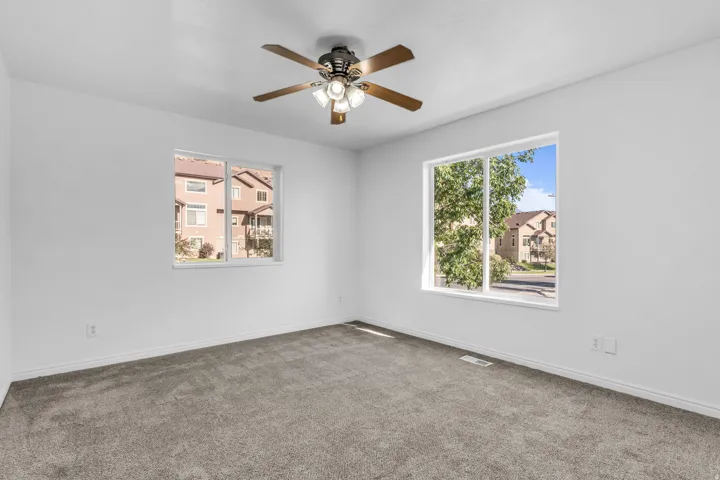 Expansive room with neutral carpeting, white walls, two large windows, and a ceiling fan with integrated lighting