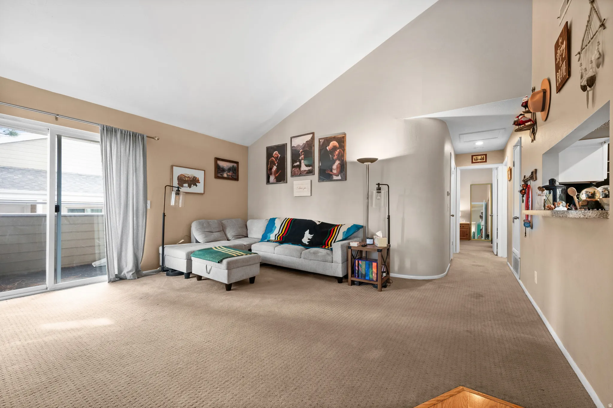 Spacious living area featuring vaulted ceilings, neutral-toned carpeting, and a sliding glass door providing access to an outdoor balcony