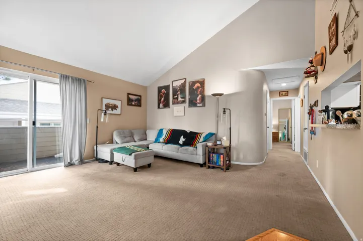 Spacious living area featuring vaulted ceilings, neutral-toned carpeting, and a sliding glass door providing access to an outdoor balcony