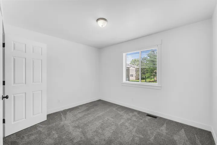 Neutral-toned room featuring a single window with horizontal blinds, gray carpeting, white walls, and a six-panel interior door with matte black hardware