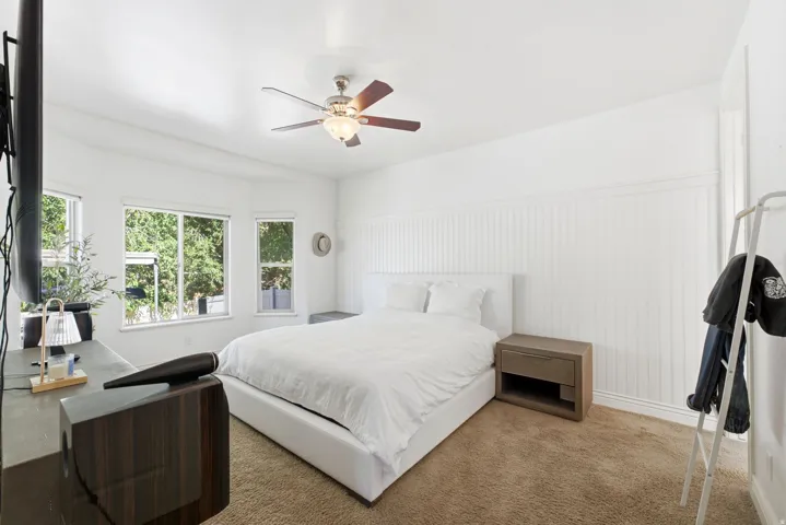Bright and airy bedroom showcasing abundant natural light from multi-pane windows, neutral white walls with vertical shiplap detailing, engineered wood-finish flooring, ceiling fan with integrated lighting, and a wood-finish nightstand