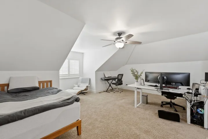 Carpeted loft space featuring sloped ceilings, a window with plantation shutters, and a ceiling fan with integrated lighting