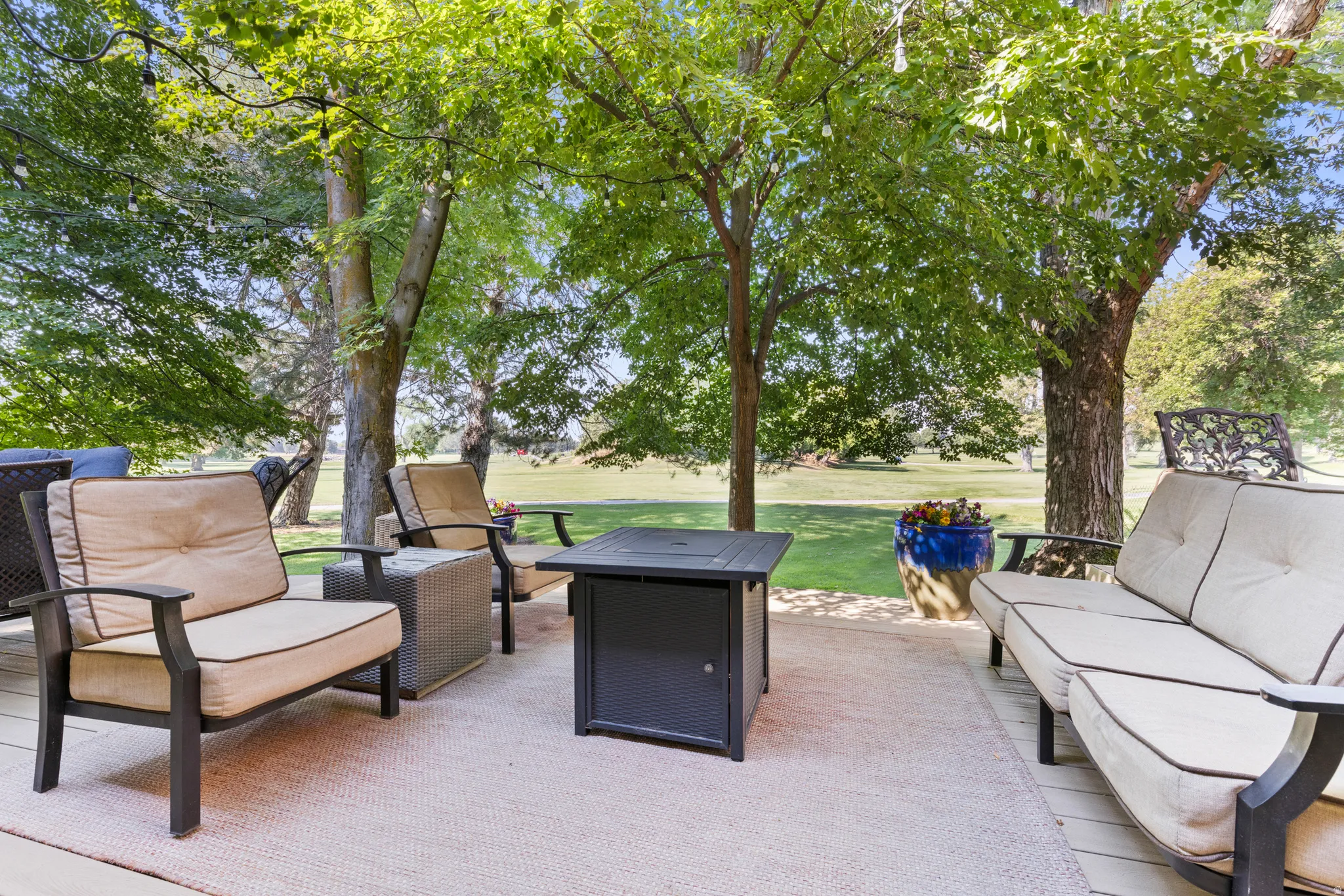 Expansive outdoor living space featuring a composite patio anchored by mature shade trees, paired seating areas with cushioned lounges, and a central fire table with a black metal base—ideal for entertaining and relaxation