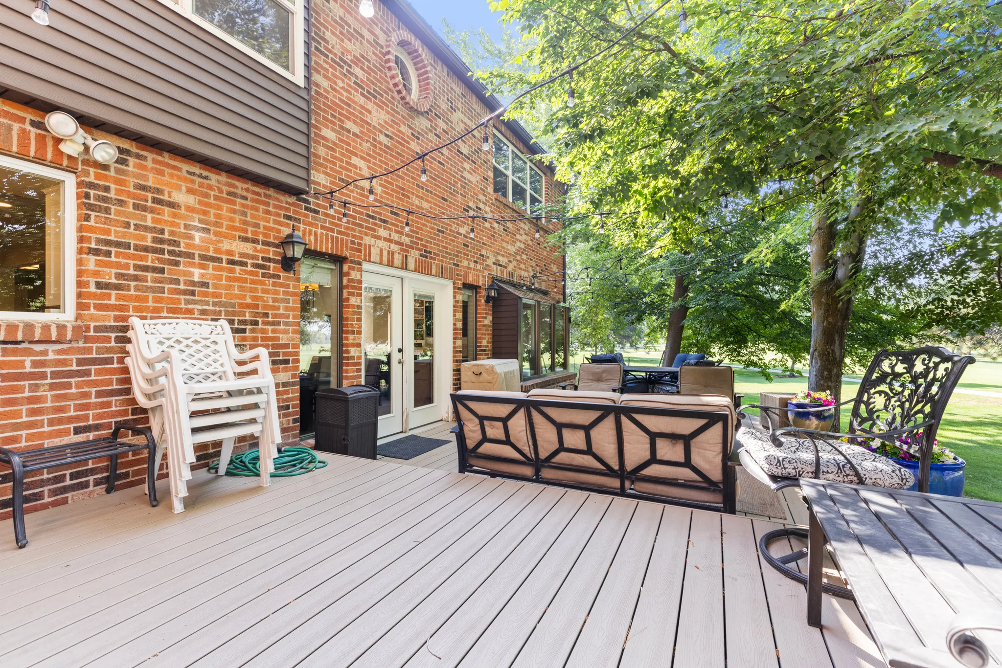 Expansive composite deck featuring gray composite decking with black metal railings and geometric panel design, anchored by a brick exterior with white trim and mature shade trees bordering the manicured lawn
