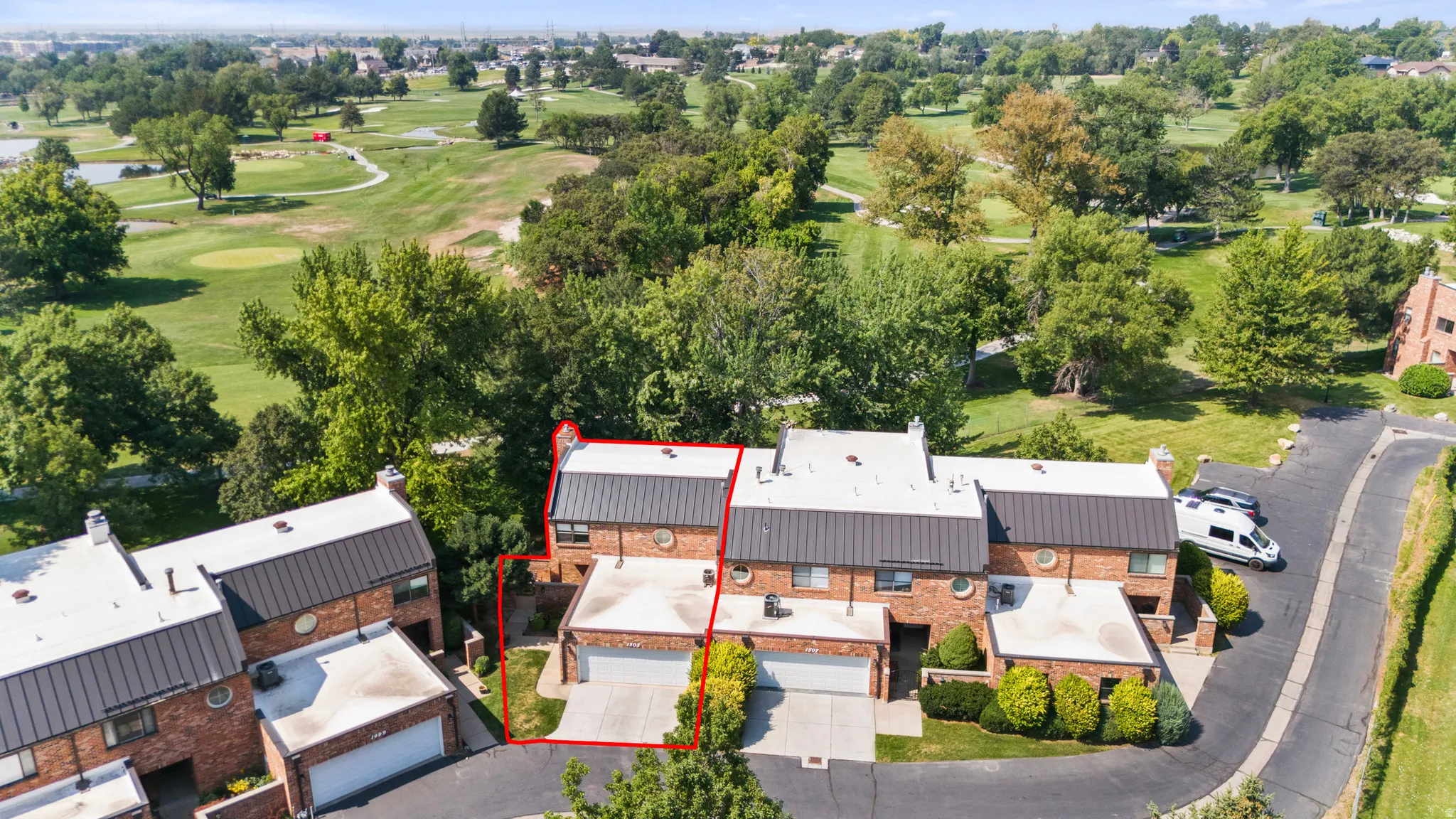 Red brick townhome with attached garage, contrasting black metal roof, and lush green landscaping