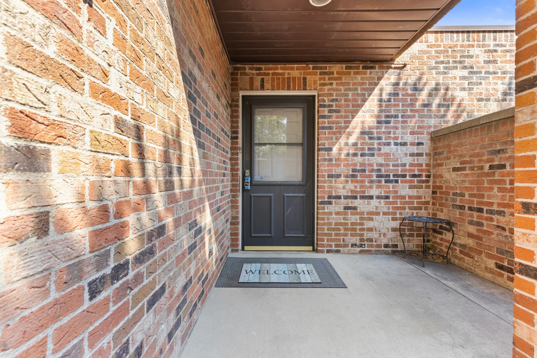 Brick exterior, concrete porch, dark-paneled entry door with screen, recessed overhead lighting, and a partial brick wall