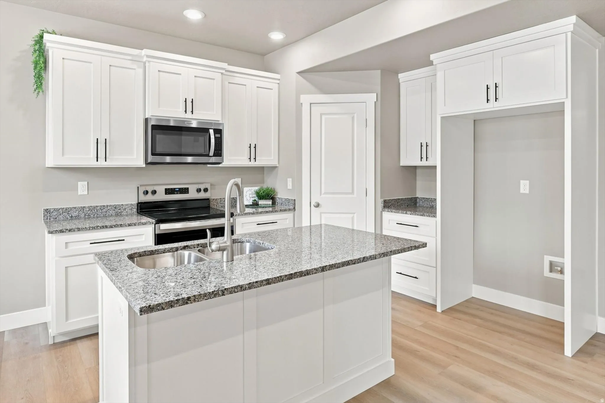 Contemporary kitchen featuring white shaker-style cabinetry, speckled stone countertops with waterfall island edge, stainless steel appliances including a gas range and microwave, chrome fixtures, and wood-finish flooring throughout the open layout