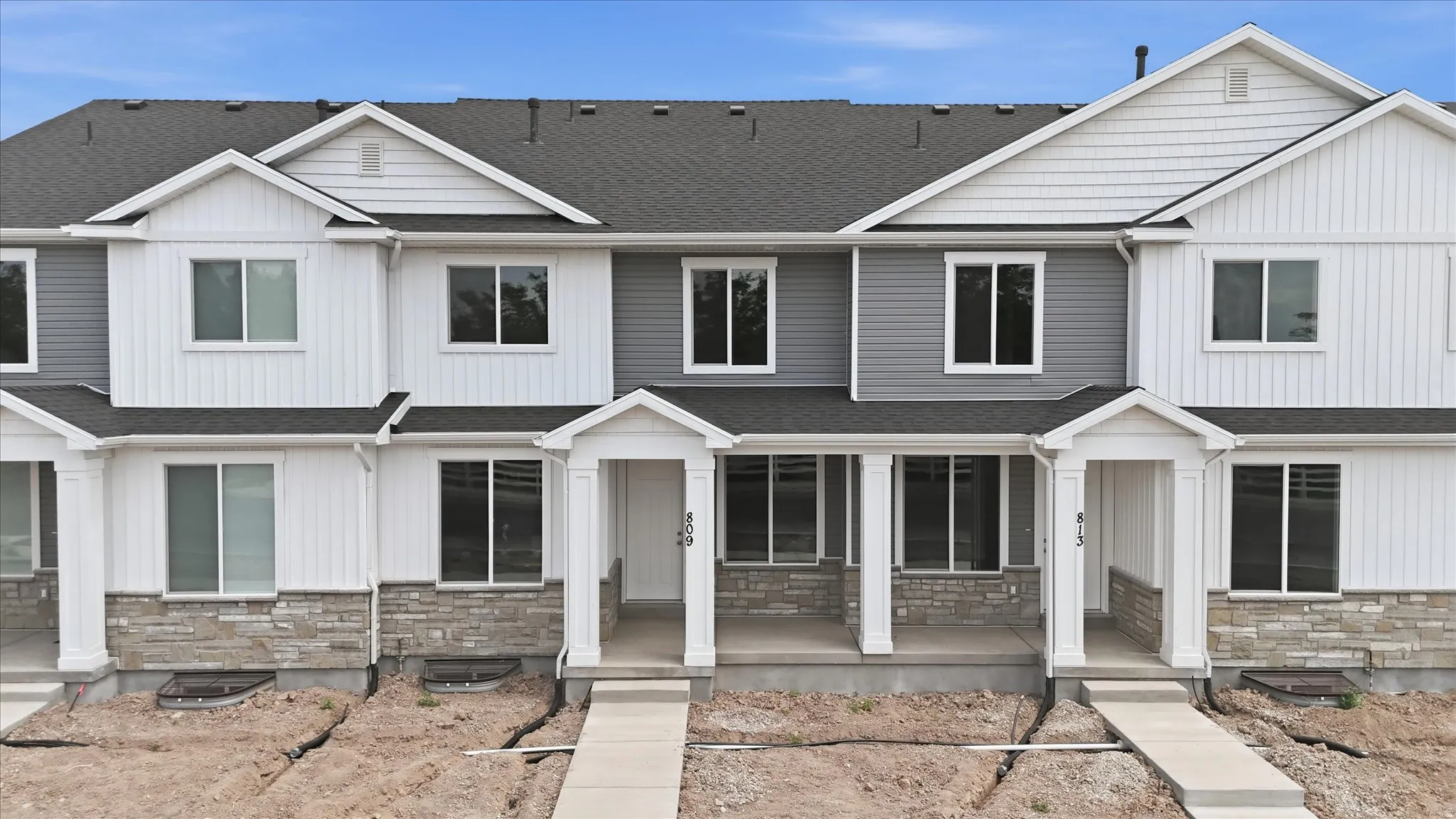 Contemporary townhome featuring clean-lined architecture with white fiber cement siding, multiple multi-pane windows, stone veneer accents on the lower facade, covered front porches with columns, and dark asphalt shingle roofing with peaked gables