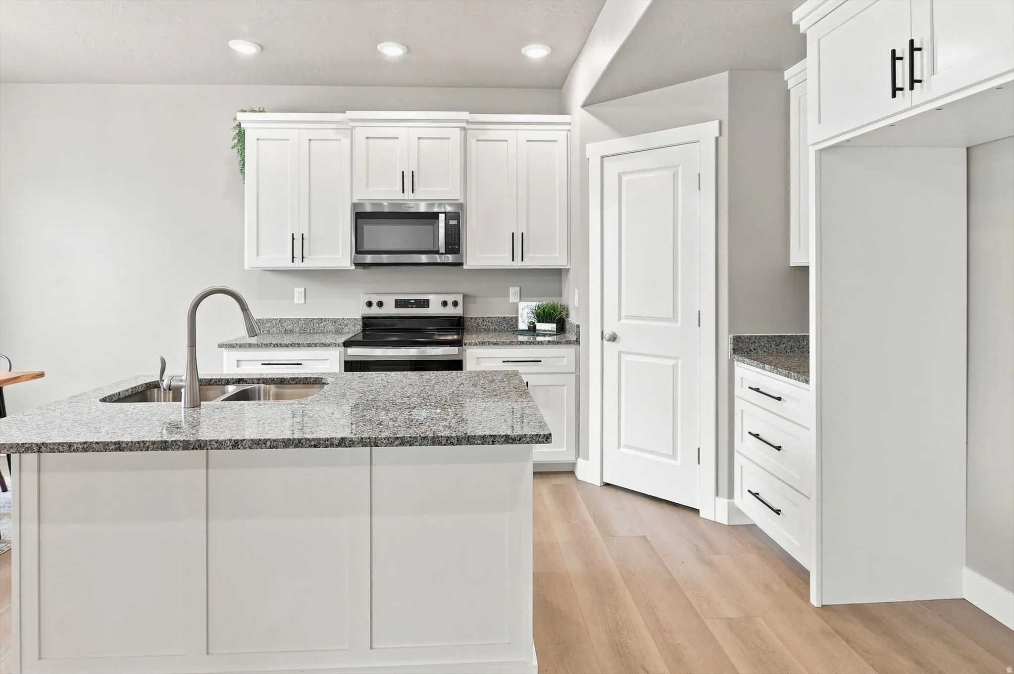 Chef's kitchen featuring white shaker-style cabinetry, stainless steel appliances, a gray quartz countertop with waterfall island edge, an arched chrome faucet, recessed lighting, and warm wood-finish flooring throughout the open layout