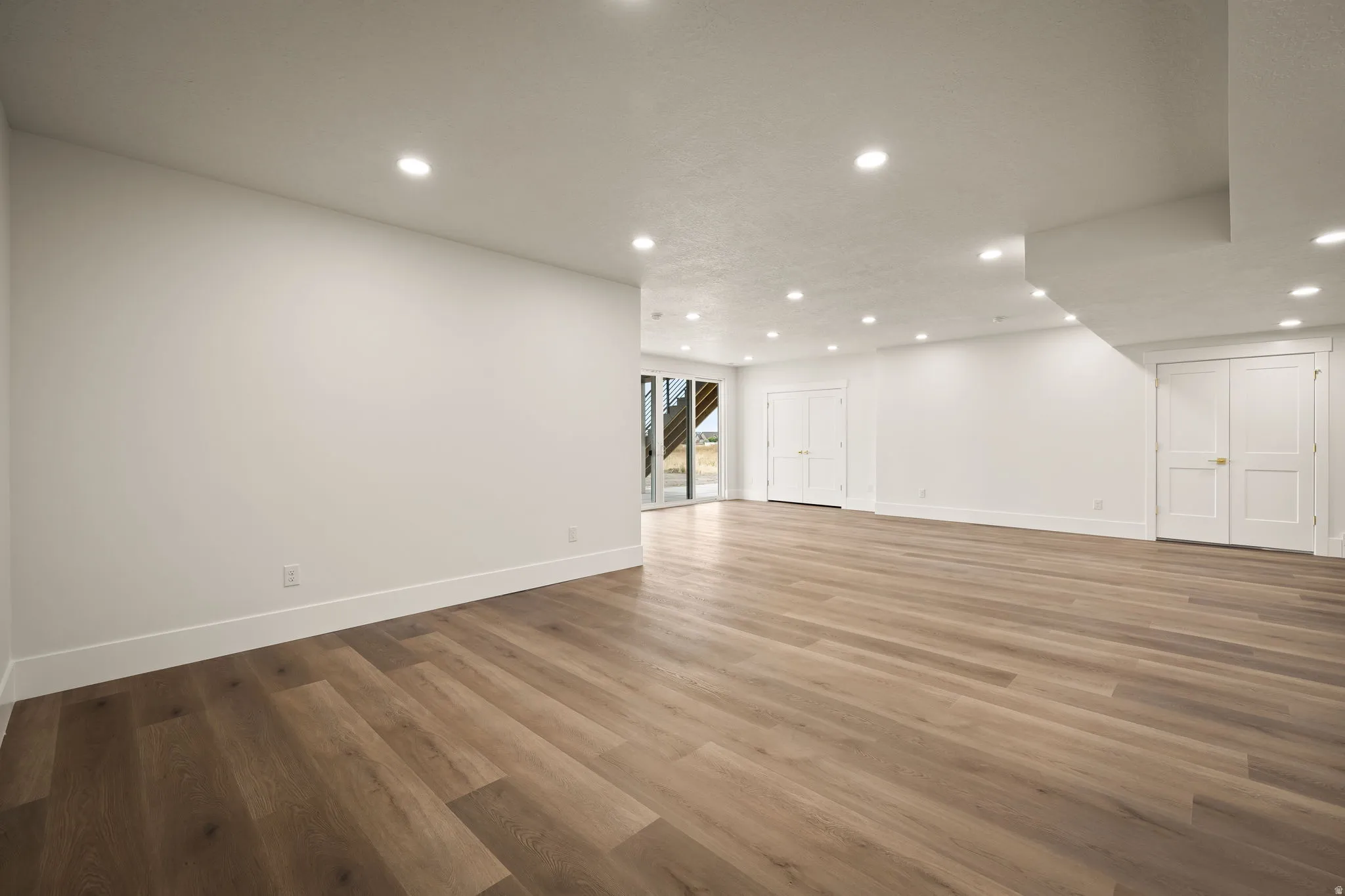 Spacious interior room featuring wood-finish flooring, recessed lighting, crisp white walls, and a large glass door providing outdoor access