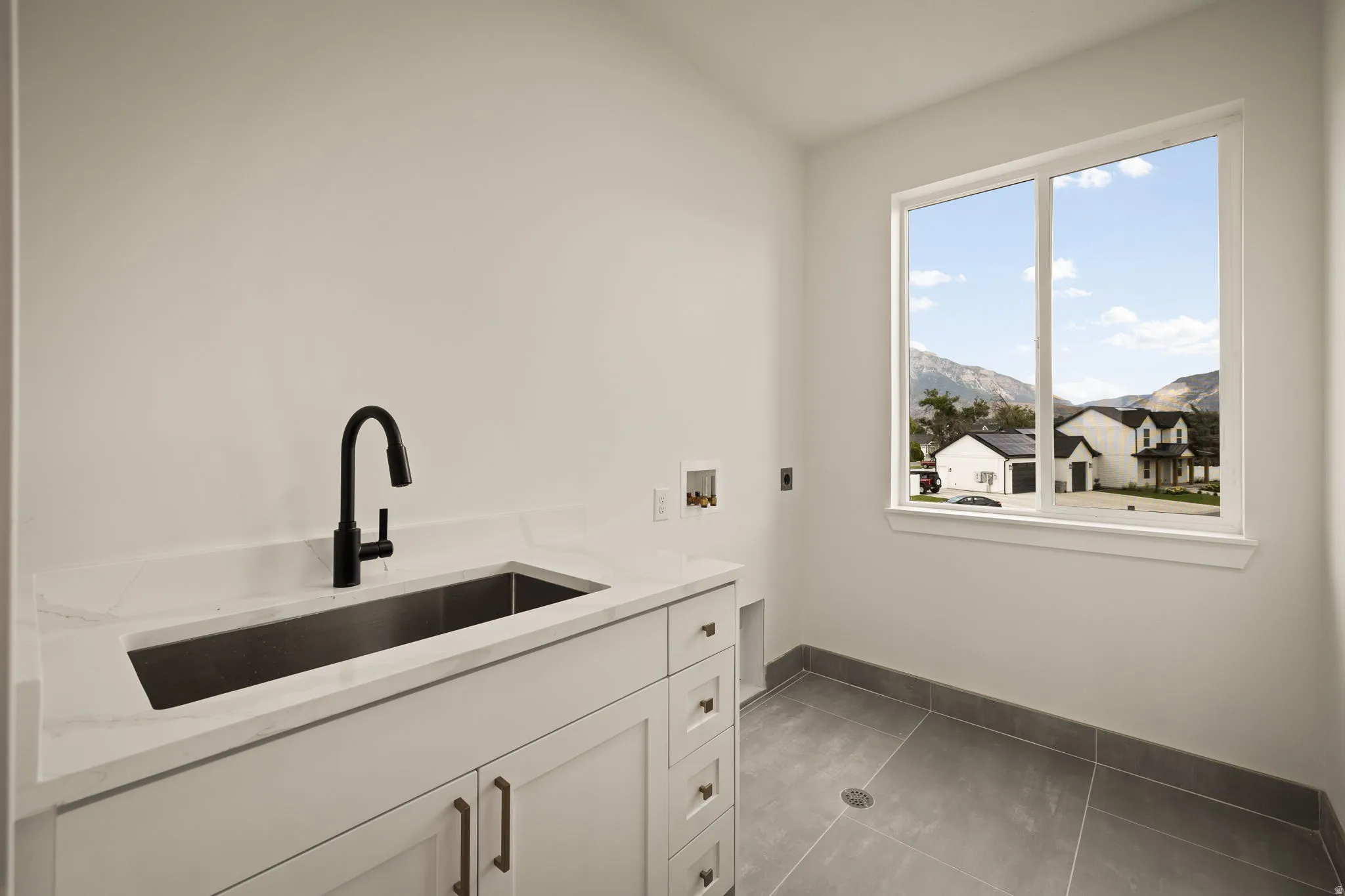 Laundry area featuring a large window with mountain scenery, a rectangular undermount sink with a matte black faucet, white shaker cabinetry with dark hardware, and gray tile flooring