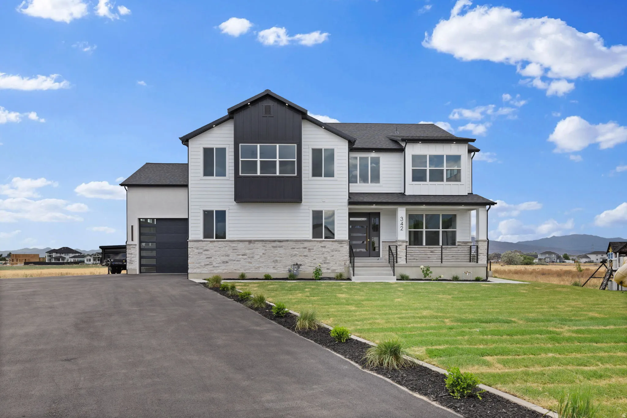 Modern two-story residence featuring white siding, dark trim accents, stone foundation elements, a covered front porch with railing, and a paved driveway