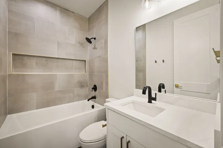 Bathroom featuring a white tub, light-toned tile surround with an integrated niche, matte black fixtures, a single vanity with a white countertop and undermount sink, and a large wall mirror