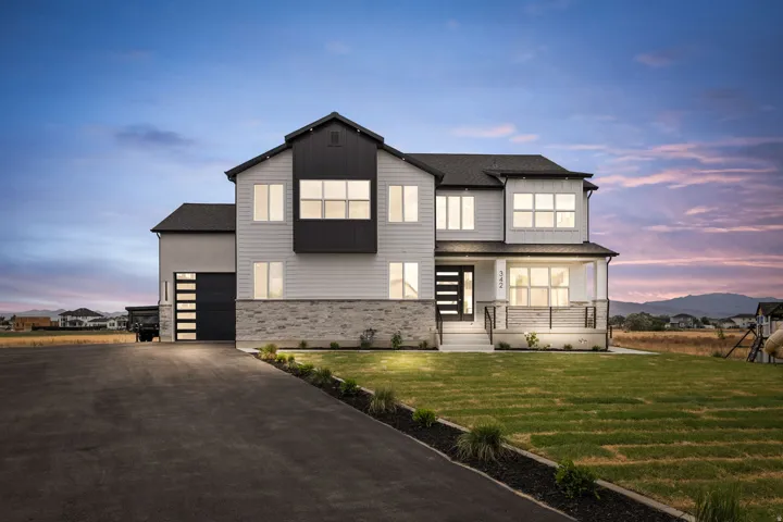 Modern two-story residence featuring a white siding exterior with dark trim accents, stone veneer detailing, a covered front porch with dark metal railings, a contemporary glass-paneled entry door, and an attached garage with a sleek panel door