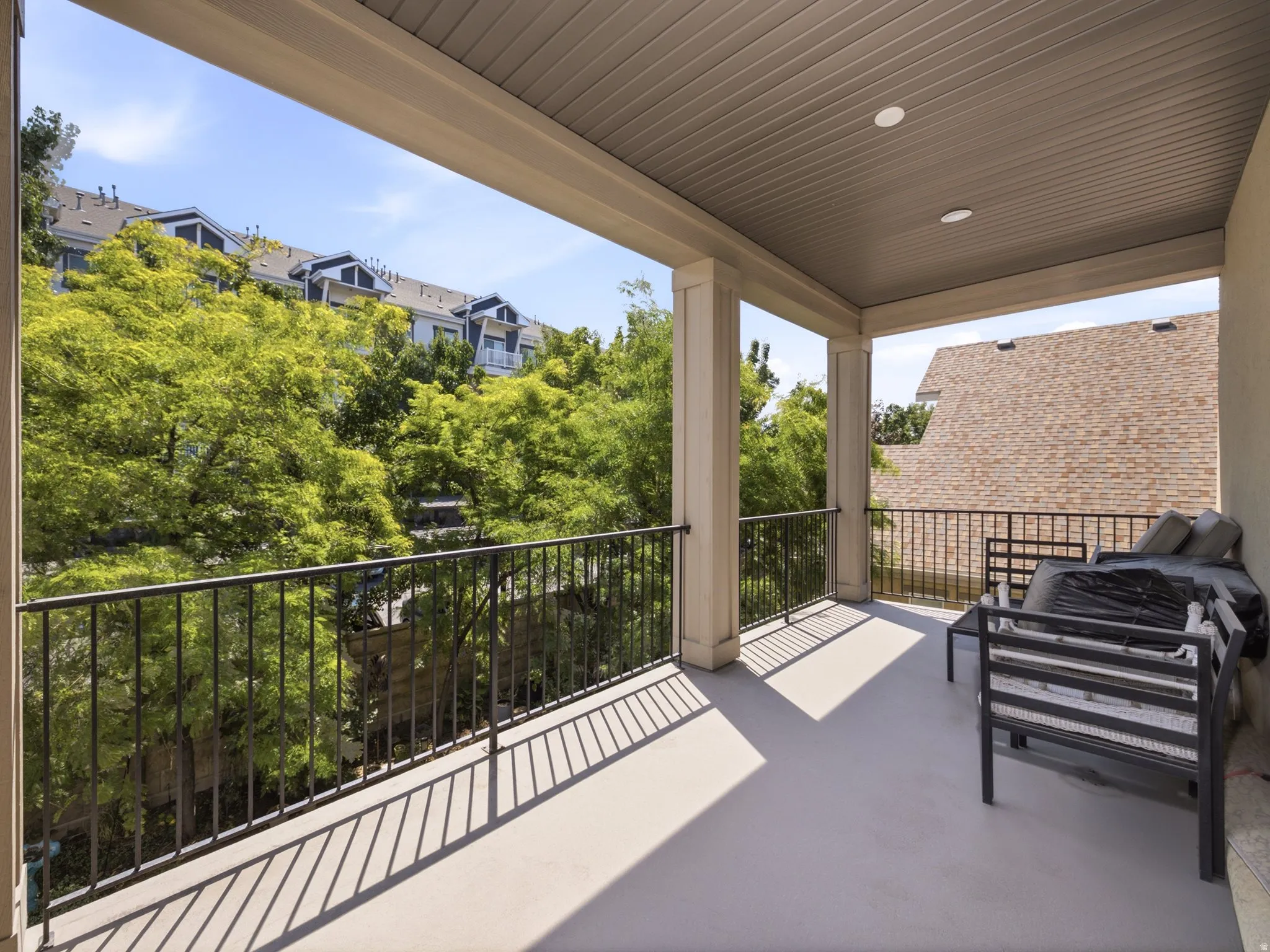 Spacious covered balcony featuring a light-toned concrete floor, solid columns, a slatted ceiling with recessed lighting, and black iron railings