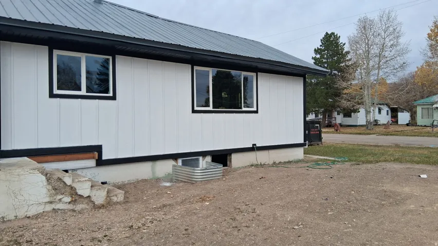 Exterior featuring white vertical siding, black trim, a dark metal roof, and a basement window well