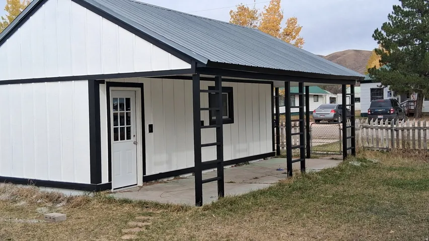 Modern farmhouse-style shed featuring a gabled metal roof, crisp white exterior paneling with contrasting black trim, and a covered front porch with black support posts