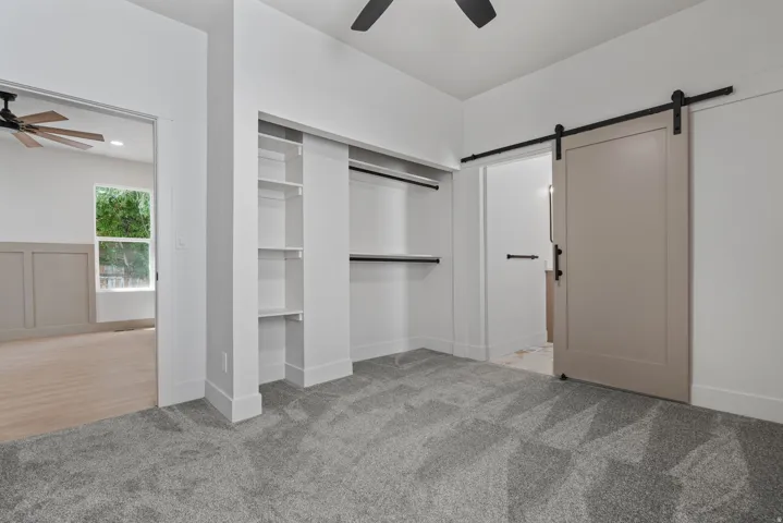 Carpeted bedroom featuring a built-in closet with shelving, a barn door entry to an adjacent room, a ceiling fan, and white baseboards