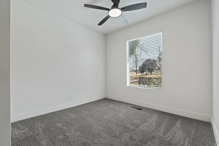 Light-filled room featuring a modern ceiling fan, a large window with horizontal blinds, and a textured gray carpet