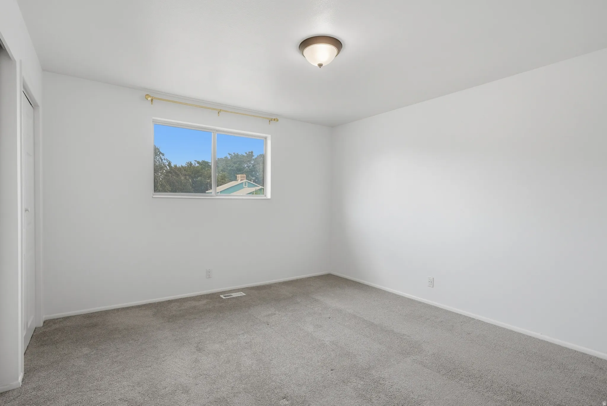Light-filled room featuring neutral wall paint, gray carpeting, a window with exterior visibility, a ceiling-mounted light fixture, and a built-in closet with white doors