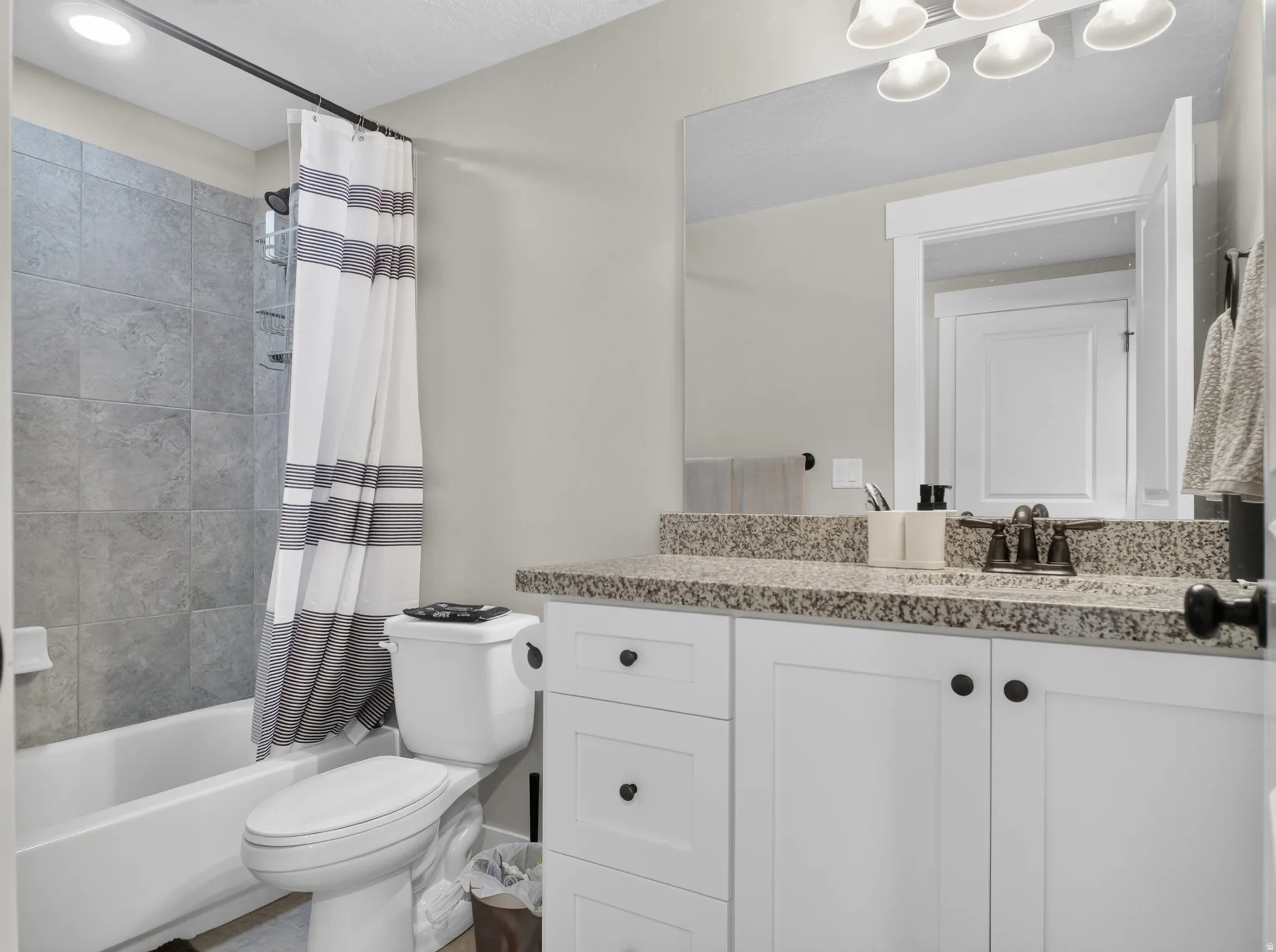 Bathroom featuring a white tub with gray tiled surround, a white vanity with a speckled countertop, a bronze-finish faucet, and a large wall-mounted mirror