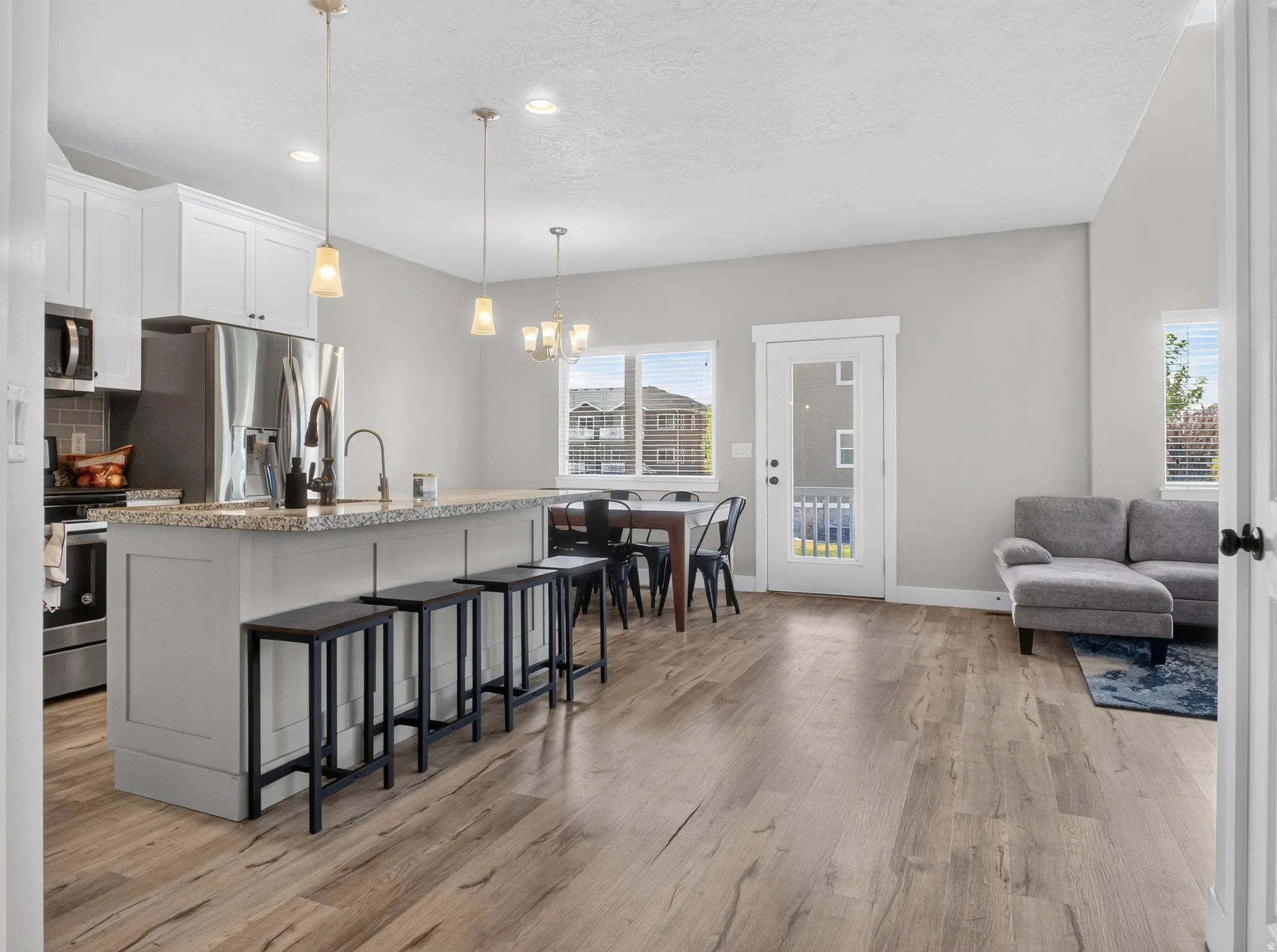 Open-concept living area featuring wood-finish flooring, a kitchen island with stone countertop, stainless steel appliances, a dining area with chandelier lighting, and a glass-paneled exterior door
