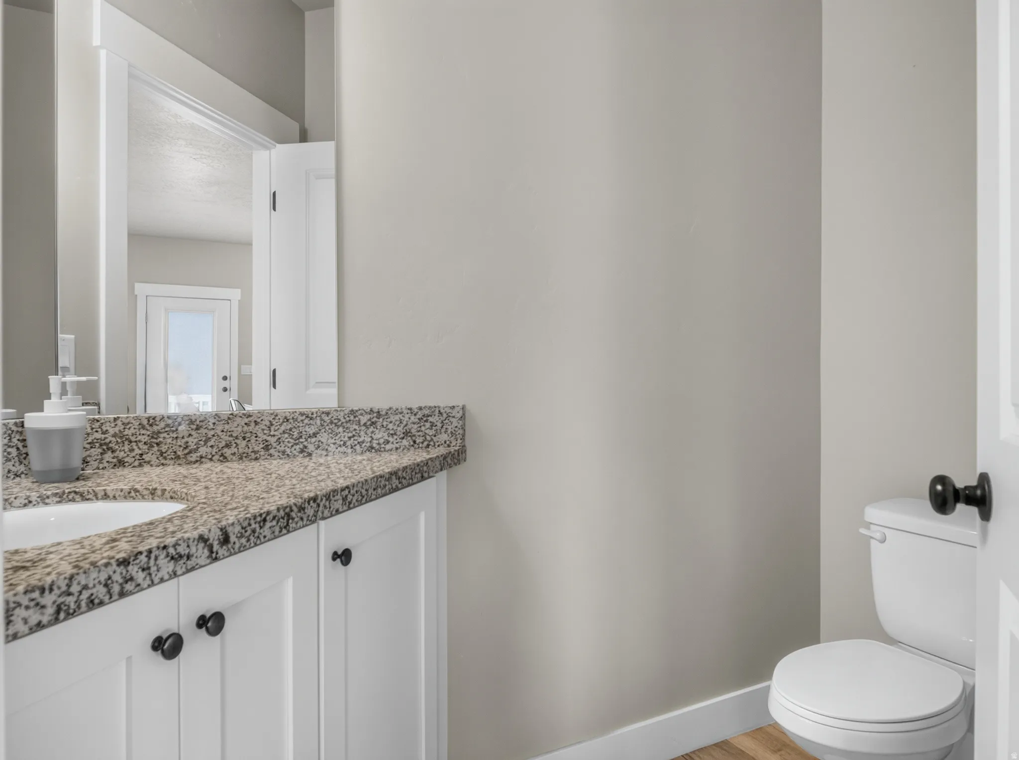 Bathroom featuring a white vanity with black hardware, a granite-finish countertop, an undermount sink, a large mirror, and wood-finish flooring